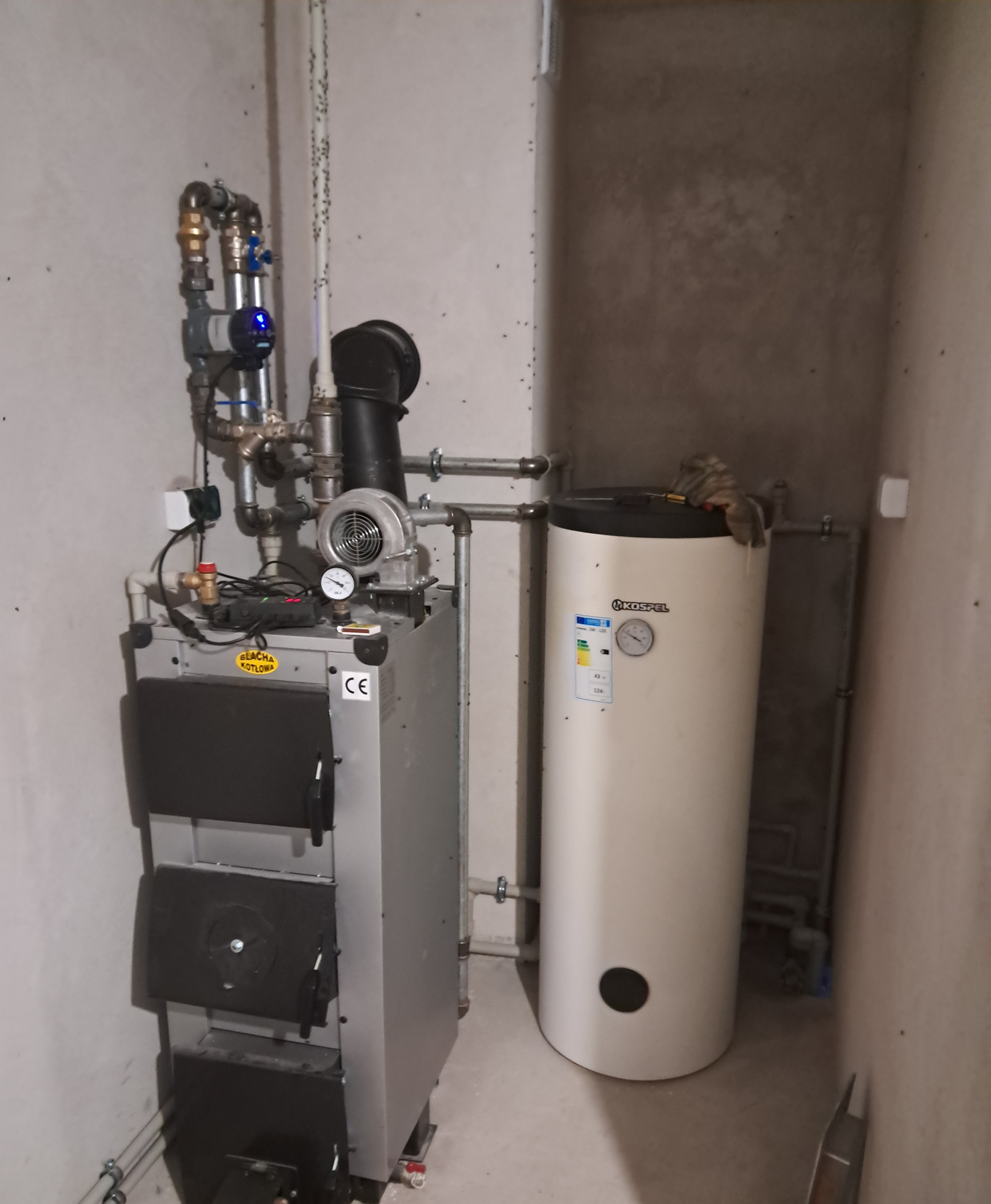 Kocioł na paliwo stałe z podajnikiem i zasobnikiem ciepłej wody użytkowej w kotłowni, widoczne rury instalacji grzewczej, manometr, wentylator i osprzęt.