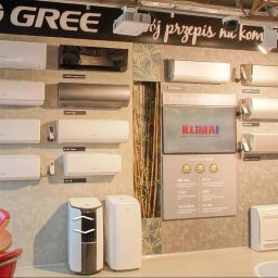 Tworzymy Instalacje Klimatyzacja Warszawa - Ekspozycja różnych modeli klimatyzatorów Gree na ścianie, w tym ścienne jednostki wewnętrzne w kolorach białym, czarnym i srebrnym, oraz przenośne klimatyzatory stojące, z widocznym logo marki...