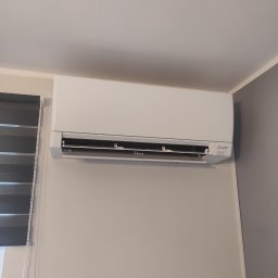 Tworzymy Instalacje Klimatyzacja Warszawa - Biała jednostka klimatyzacyjna Mitsubishi Electric zamontowana na ścianie w pokoju z szarymi roletami okiennymi.