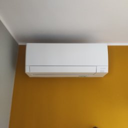 Tworzymy Instalacje Klimatyzacja Warszawa - Biała jednostka klimatyzacji Mitsubishi Electric zamontowana na musztardowej ścianie w pomieszczeniu z białym sufitem i fragmentem szarej ściany, widoczny fragment czarnej dekoracji na ścianie.