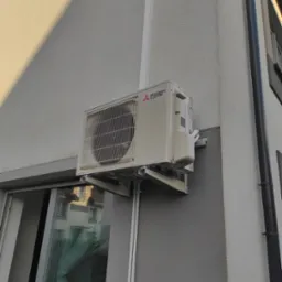 Zewnętrzna jednostka klimatyzatora Mitsubishi Electric zamontowana na ścianie budynku z szarą elewacją, widoczna z boku, z odbiciem otoczenia w oknie poniżej.