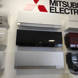 Tworzymy Instalacje Klimatyzacja Warszawa - Wystawa modeli klimatyzatorów Mitsubishi Electric w różnych kolorach (biały, czarny, srebrny, bordowy) zamontowanych na białej ścianie pod logo producenta.