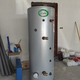 Tworzymy Instalacje Klimatyzacja Warszawa - Szary, cylindryczny zasobnik ciepłej wody użytkowej marki Joule, z widocznymi przyłączami i logo, ustawiony w surowym wnętrzu w trakcie montażu instalacji grzewczej.