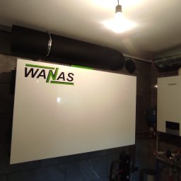 Tworzymy Instalacje Klimatyzacja Warszawa - Biała centrala wentylacyjna z rekuperacją marki WANAS zamontowana na ścianie z szarych płytek imitujących marmur, widoczne czarne rury wentylacyjne i piec gazowy Vaillant.