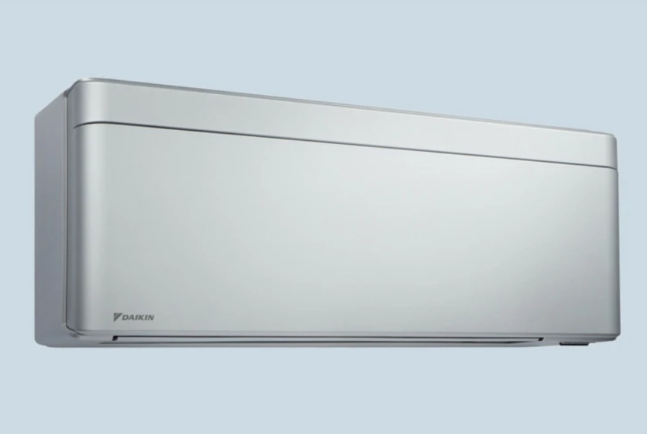 Elegancki, srebrny klimatyzator ścienny marki Daikin na jednolitym, jasnoniebieskim tle, prezentujący minimalistyczny design z subtelnymi zaokrągleniami.