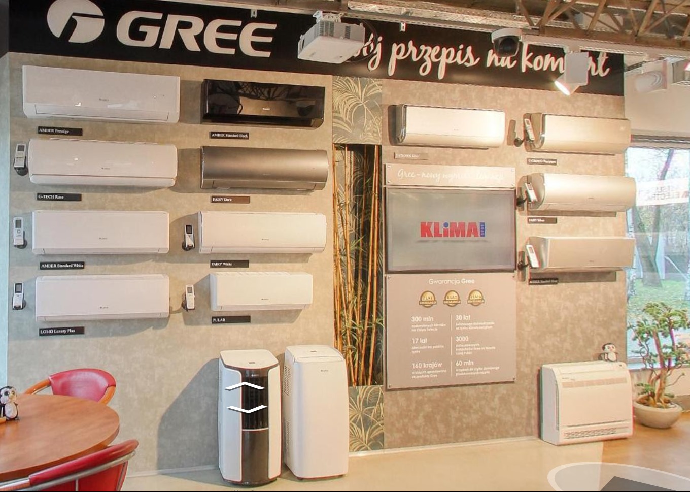 Ekspozycja różnych modeli klimatyzatorów Gree na ścianie, w tym ścienne jednostki wewnętrzne w kolorach białym, czarnym i srebrnym, oraz przenośne klimatyzatory stojące, z widocznym logo marki...
