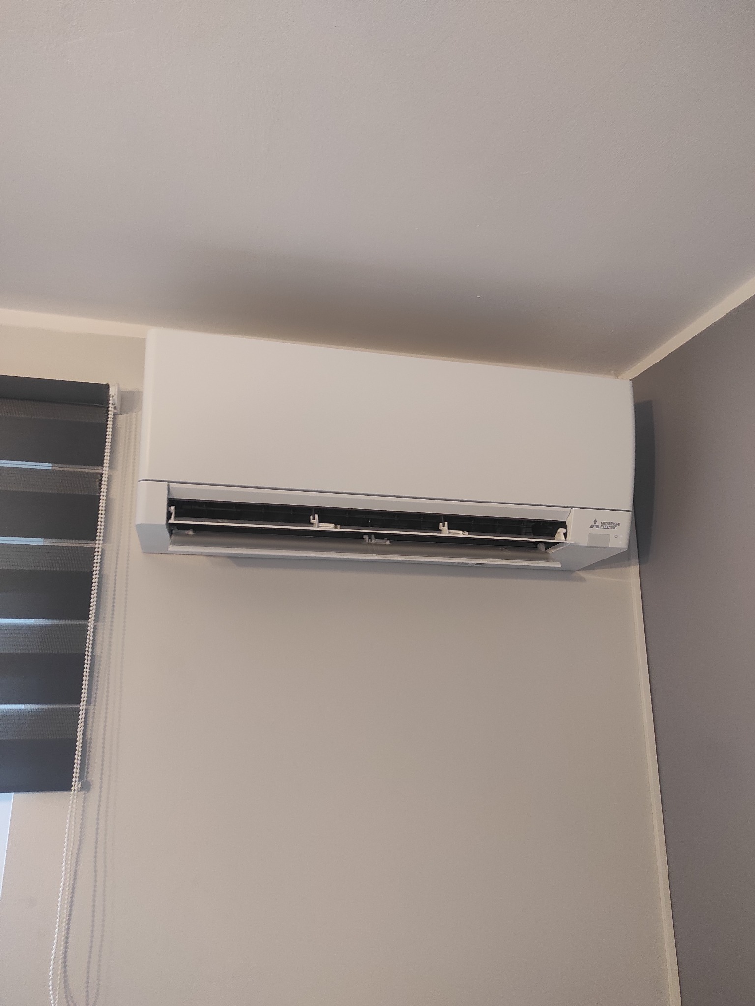 Biała jednostka klimatyzacyjna Mitsubishi Electric zamontowana na ścianie w pokoju z szarymi roletami okiennymi.