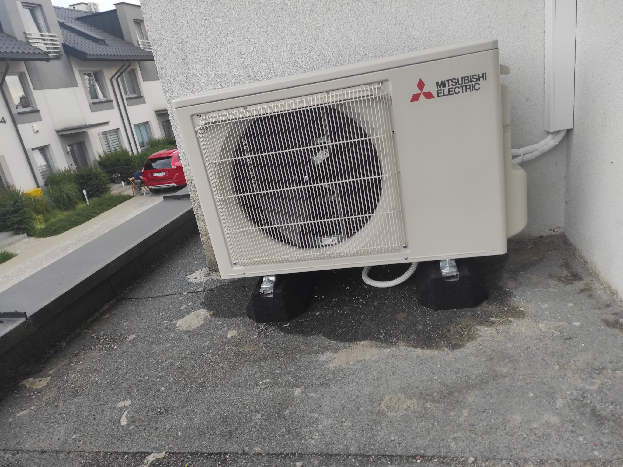 Zewnętrzna jednostka klimatyzacji Mitsubishi Electric zamontowana na dachu budynku mieszkalnego, widoczne czarne podkładki antywibracyjne i białe rury odprowadzające skropliny.
