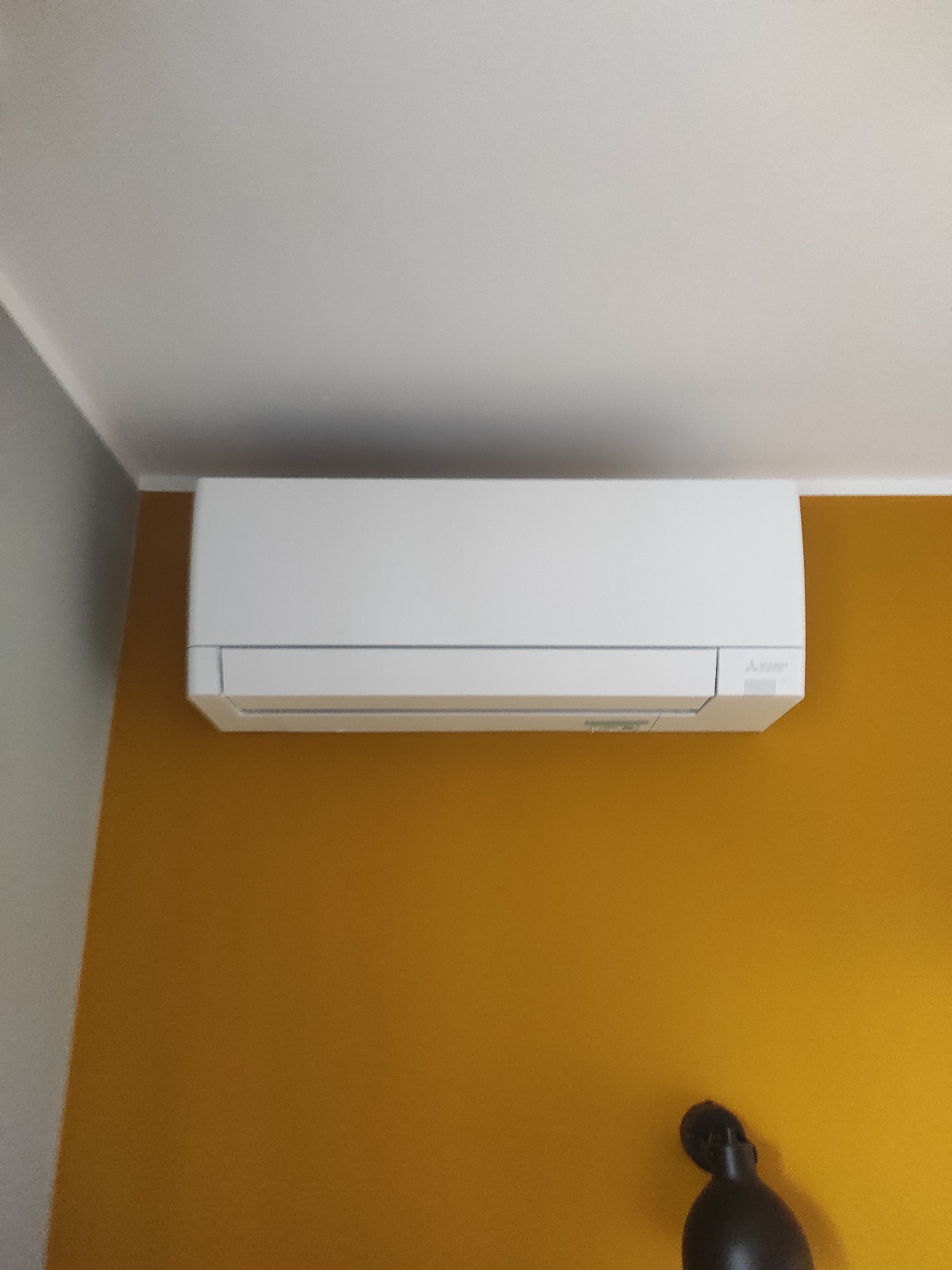 Biała jednostka klimatyzacji Mitsubishi Electric zamontowana na musztardowej ścianie w pomieszczeniu z białym sufitem i fragmentem szarej ściany, widoczny fragment czarnej dekoracji na ścianie.