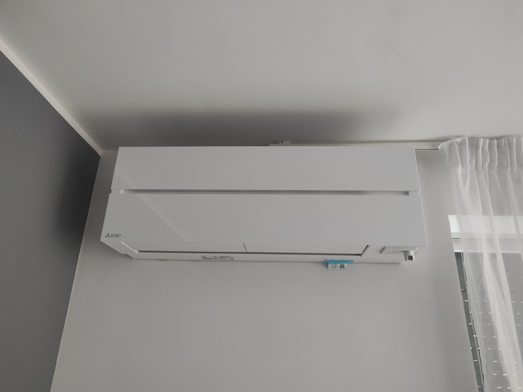 Biała jednostka klimatyzacji Mitsubishi Electric zamontowana na szarej ścianie w pomieszczeniu, widoczna firanka w oknie po prawej stronie.