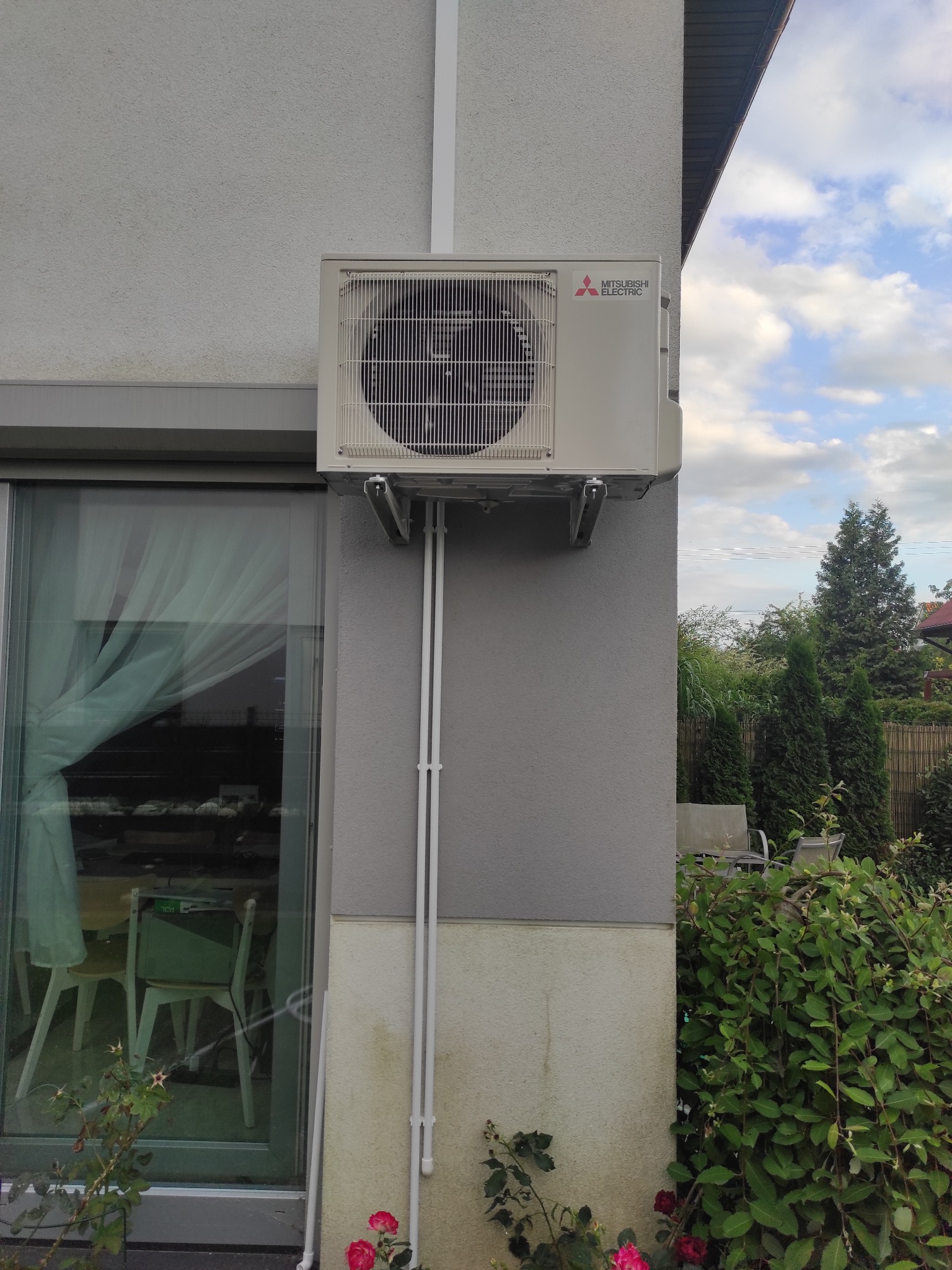 Zewnętrzna jednostka klimatyzacji Mitsubishi Electric zamontowana na szarej ścianie domu z widocznymi białymi rurami instalacyjnymi, z fragmentem okna i zielenią w tle.