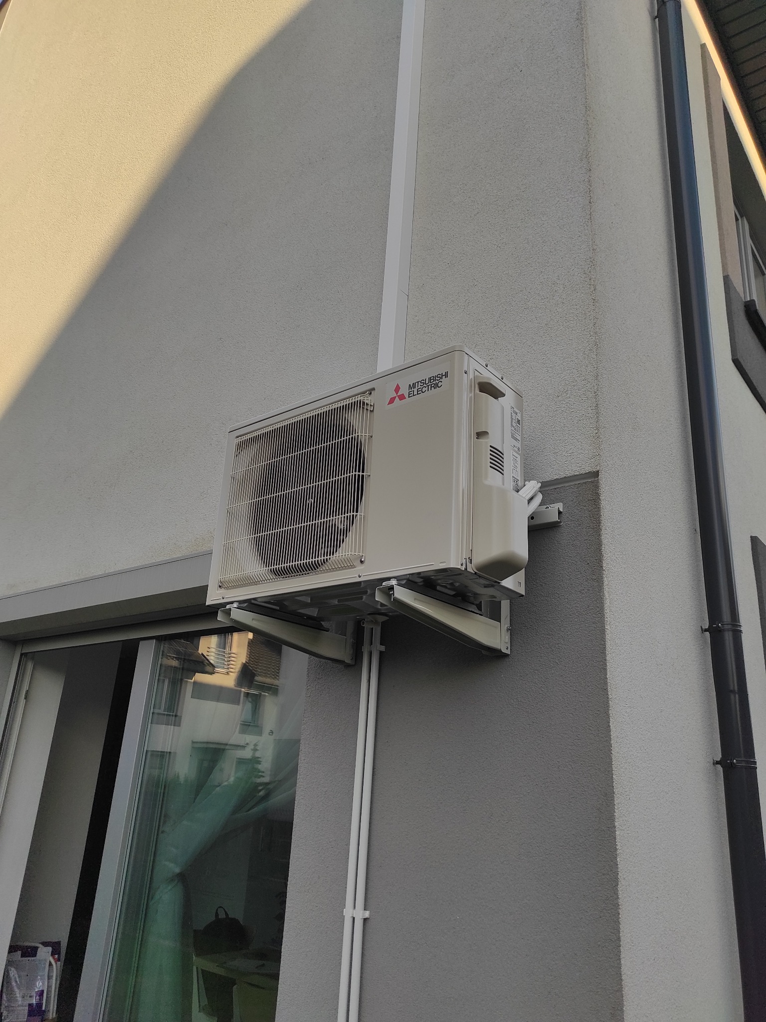 Zewnętrzna jednostka klimatyzatora Mitsubishi Electric zamontowana na ścianie budynku z szarą elewacją, widoczna z boku, z odbiciem otoczenia w oknie poniżej.
