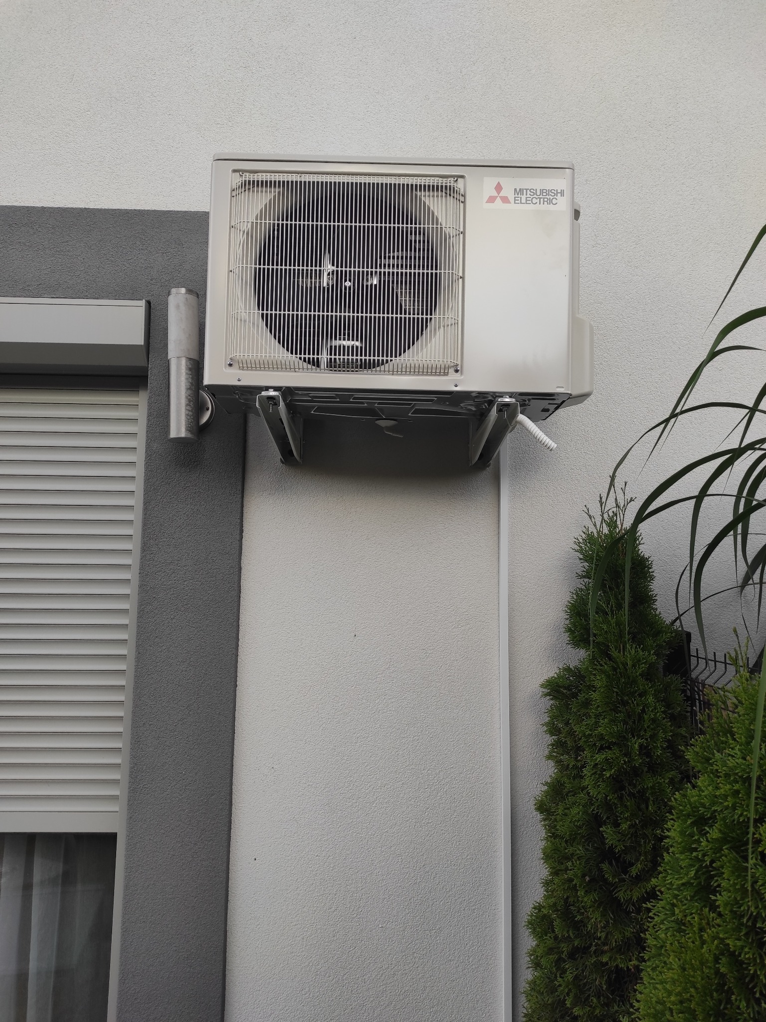 Zewnętrzna jednostka klimatyzacji Mitsubishi Electric zamontowana na jasnej ścianie budynku, widoczne szare elementy elewacji, roleta okienna oraz iglaki w tle.
