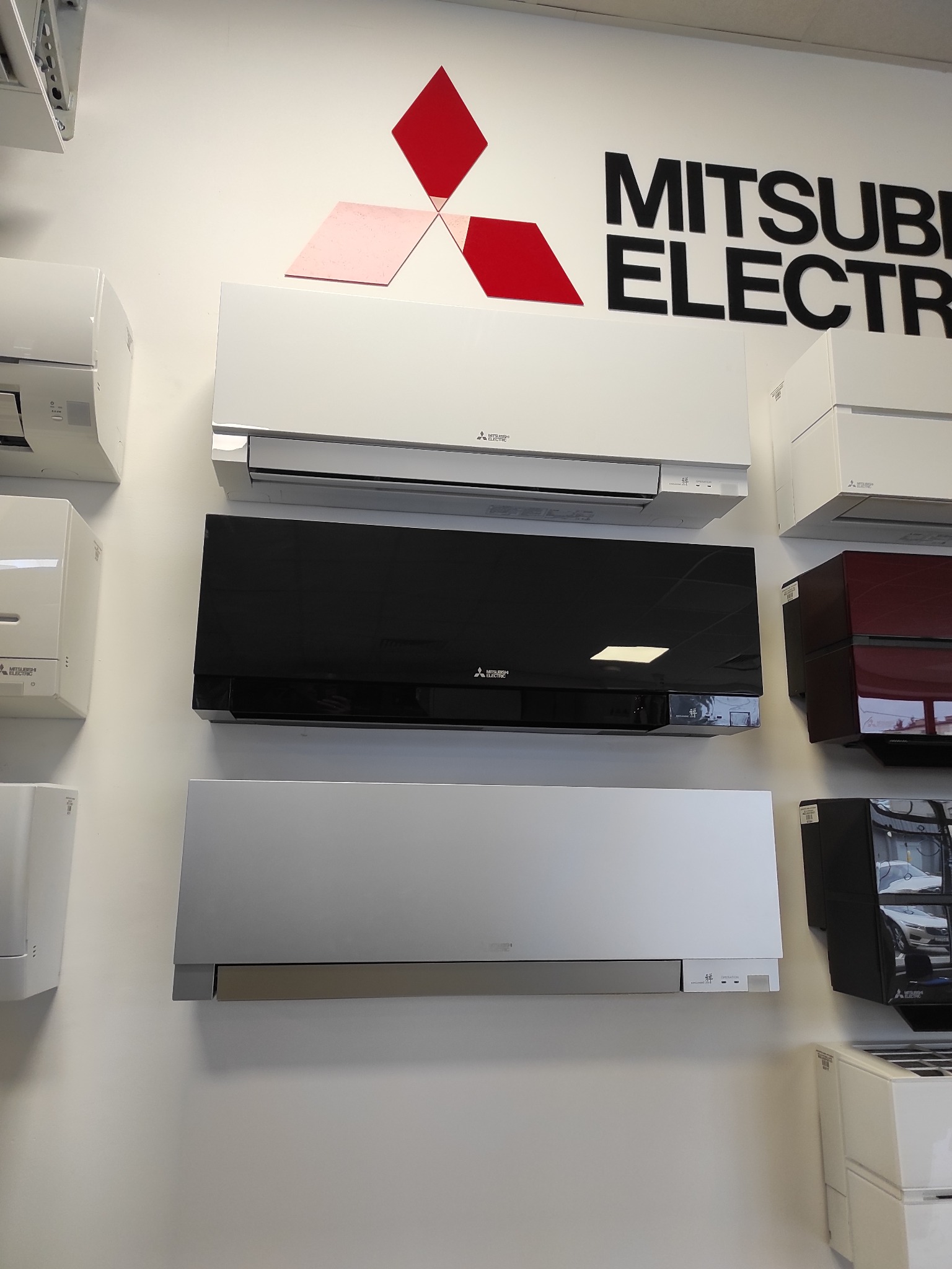 Wystawa modeli klimatyzatorów Mitsubishi Electric w różnych kolorach (biały, czarny, srebrny, bordowy) zamontowanych na białej ścianie pod logo producenta.