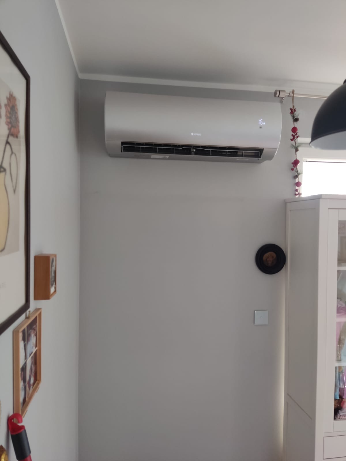 Zamontowany na ścianie, szary klimatyzator marki Gree w jasnym pomieszczeniu mieszkalnym z dekoracjami i ramkami na ścianie.