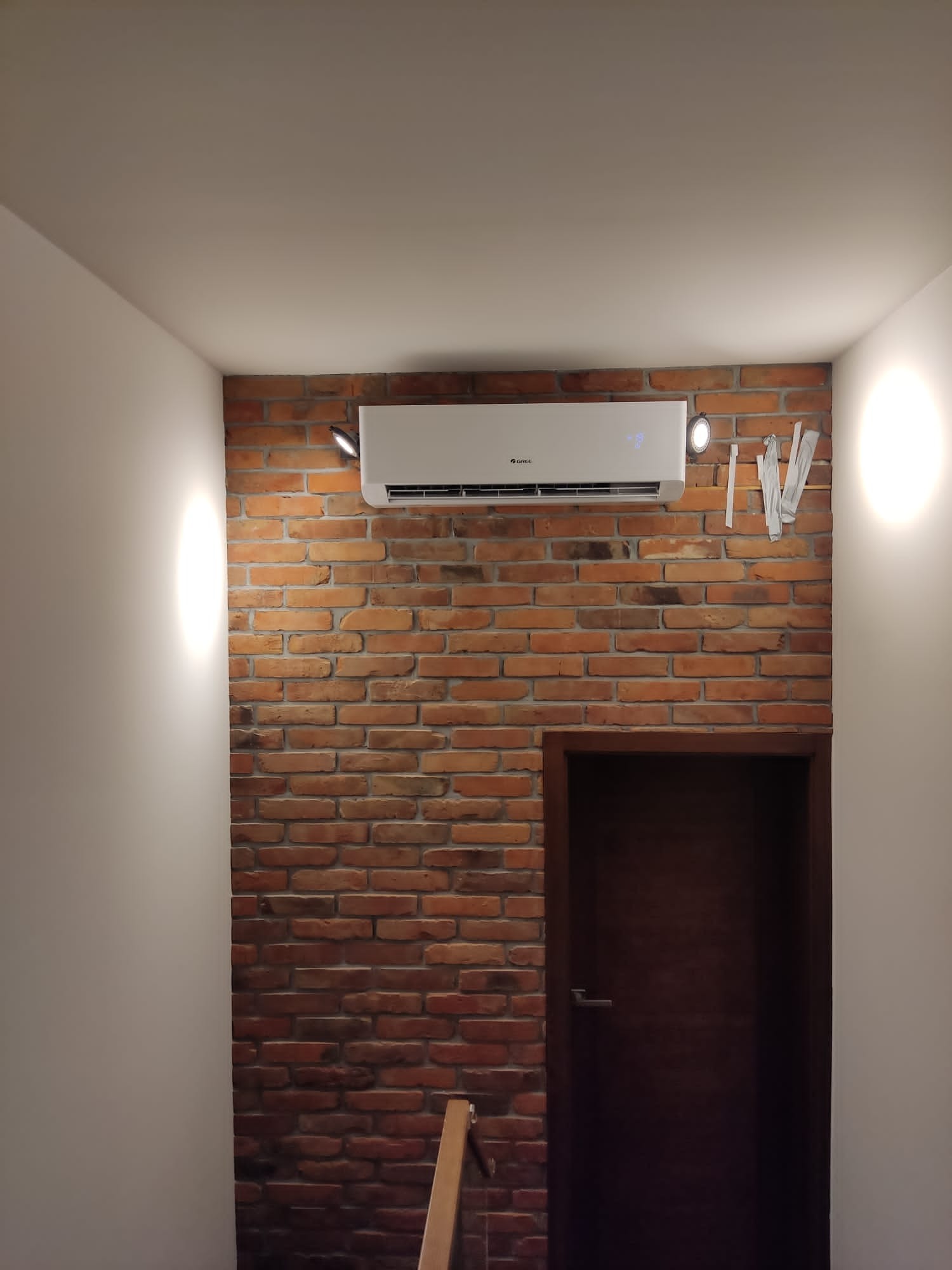 Biały klimatyzator marki Gree zamontowany na ścianie z czerwonej cegły w pomieszczeniu z ciemnymi drzwiami i oświetleniem punktowym.