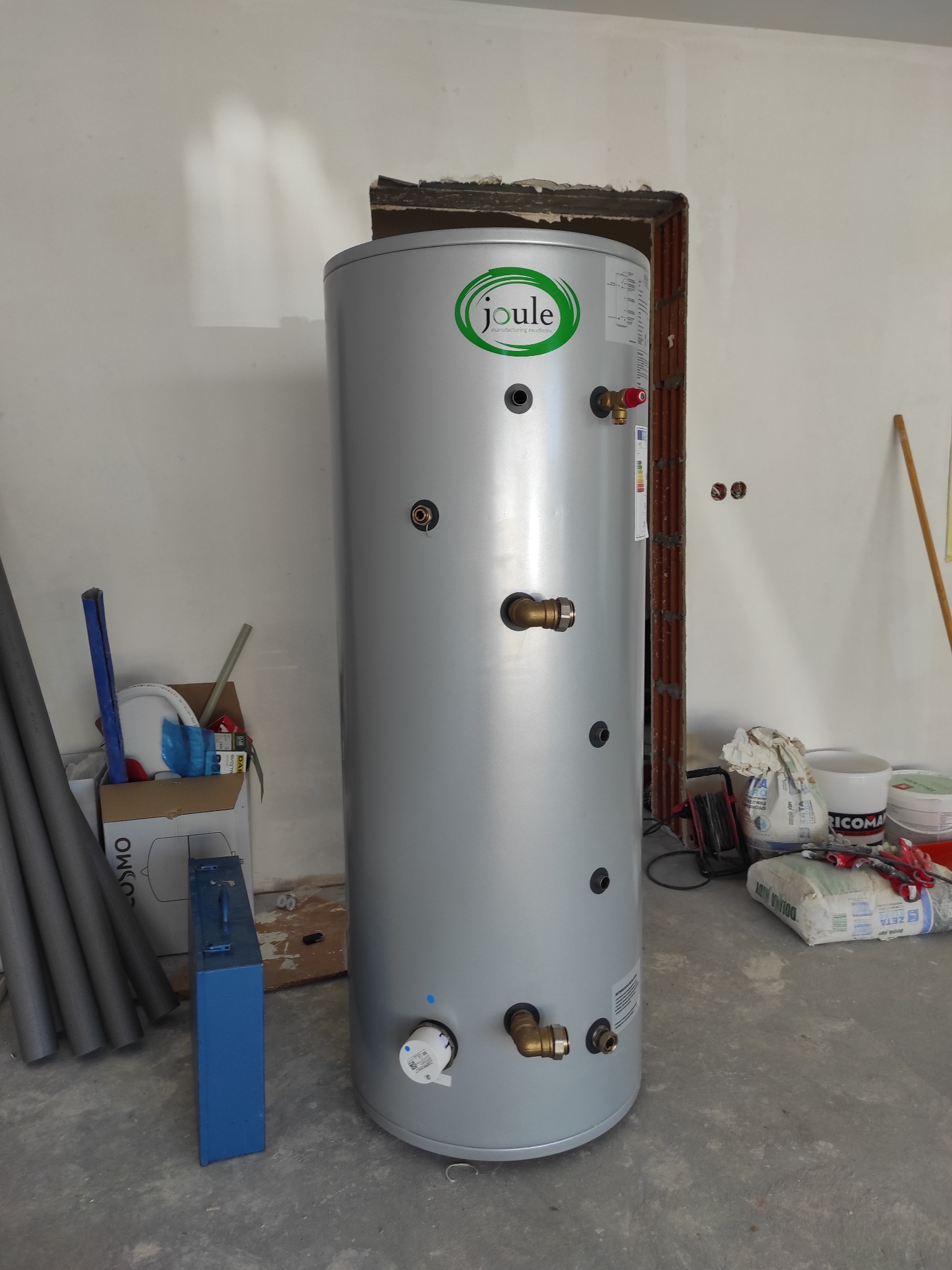 Szary, cylindryczny zasobnik ciepłej wody użytkowej marki Joule, z widocznymi przyłączami i logo, ustawiony w surowym wnętrzu w trakcie montażu instalacji grzewczej.