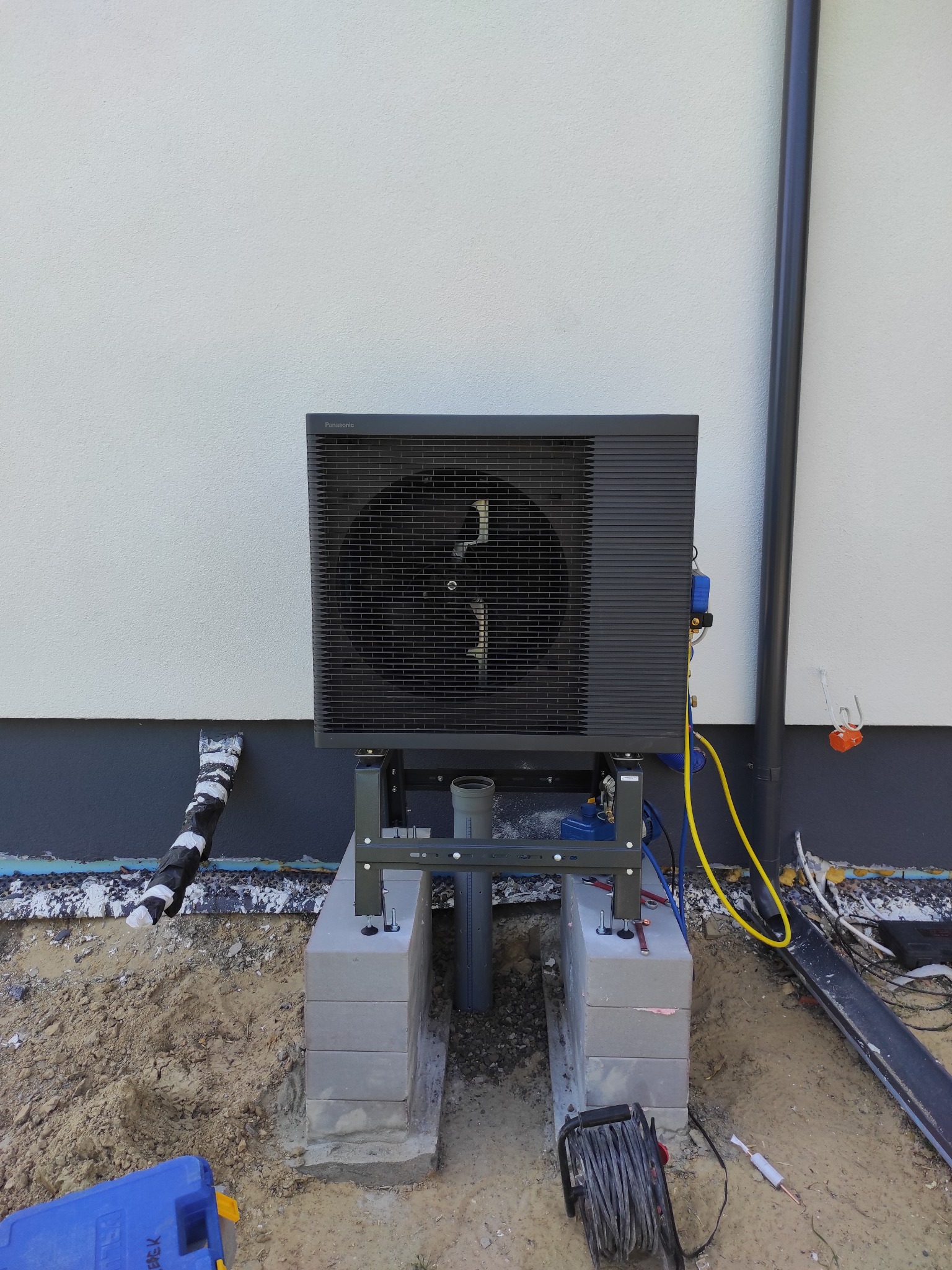 Szara pompa ciepła Panasonic zamontowana na betonowych bloczkach przy ścianie budynku z widocznymi elementami instalacji i narzędziami.
