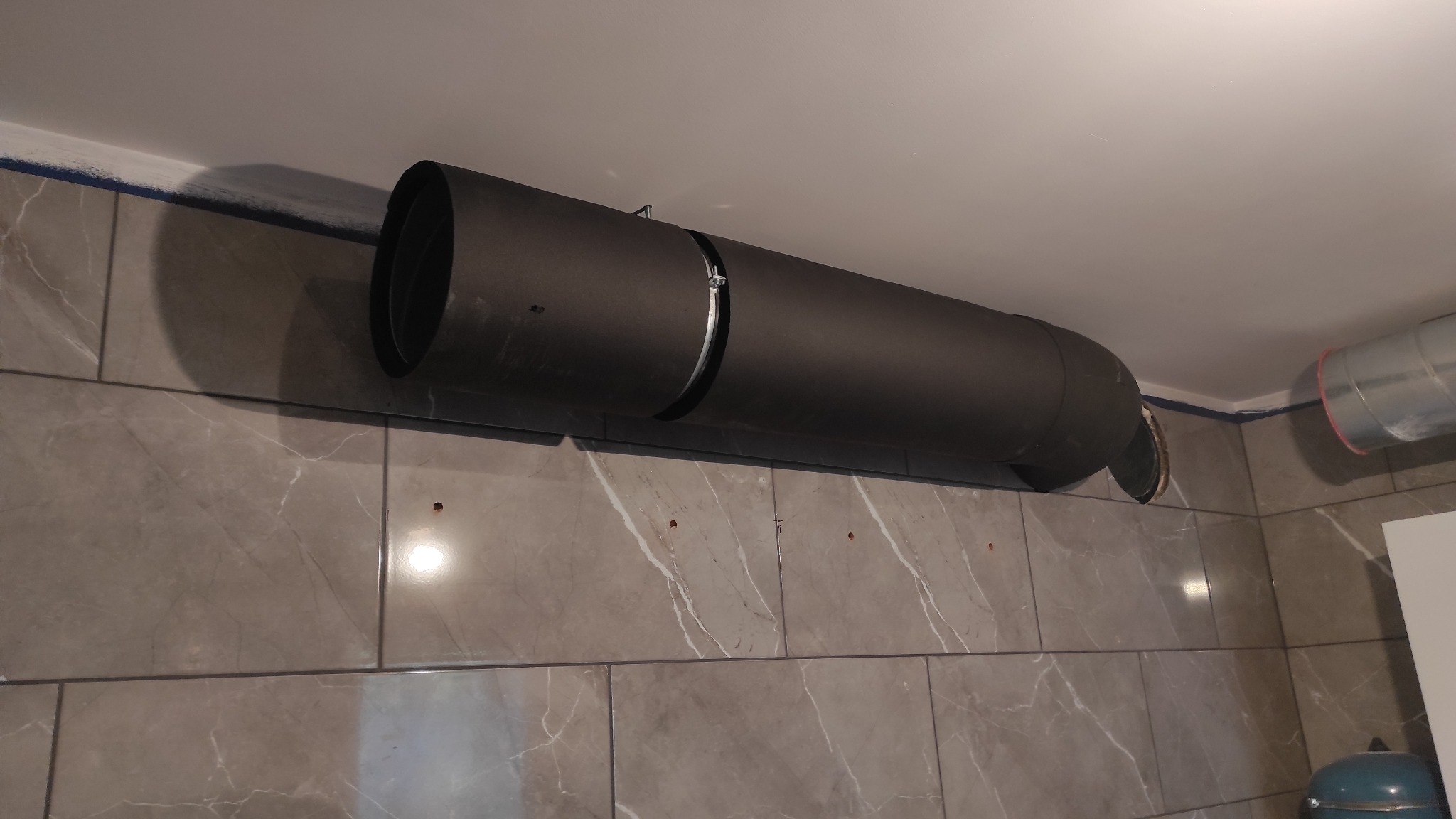 Czarny kanał wentylacyjny rekuperacji, zamocowany do sufitu i ściany wyłożonej płytkami imitującymi marmur, widoczny łącznik kanałów z metalową obejmą.