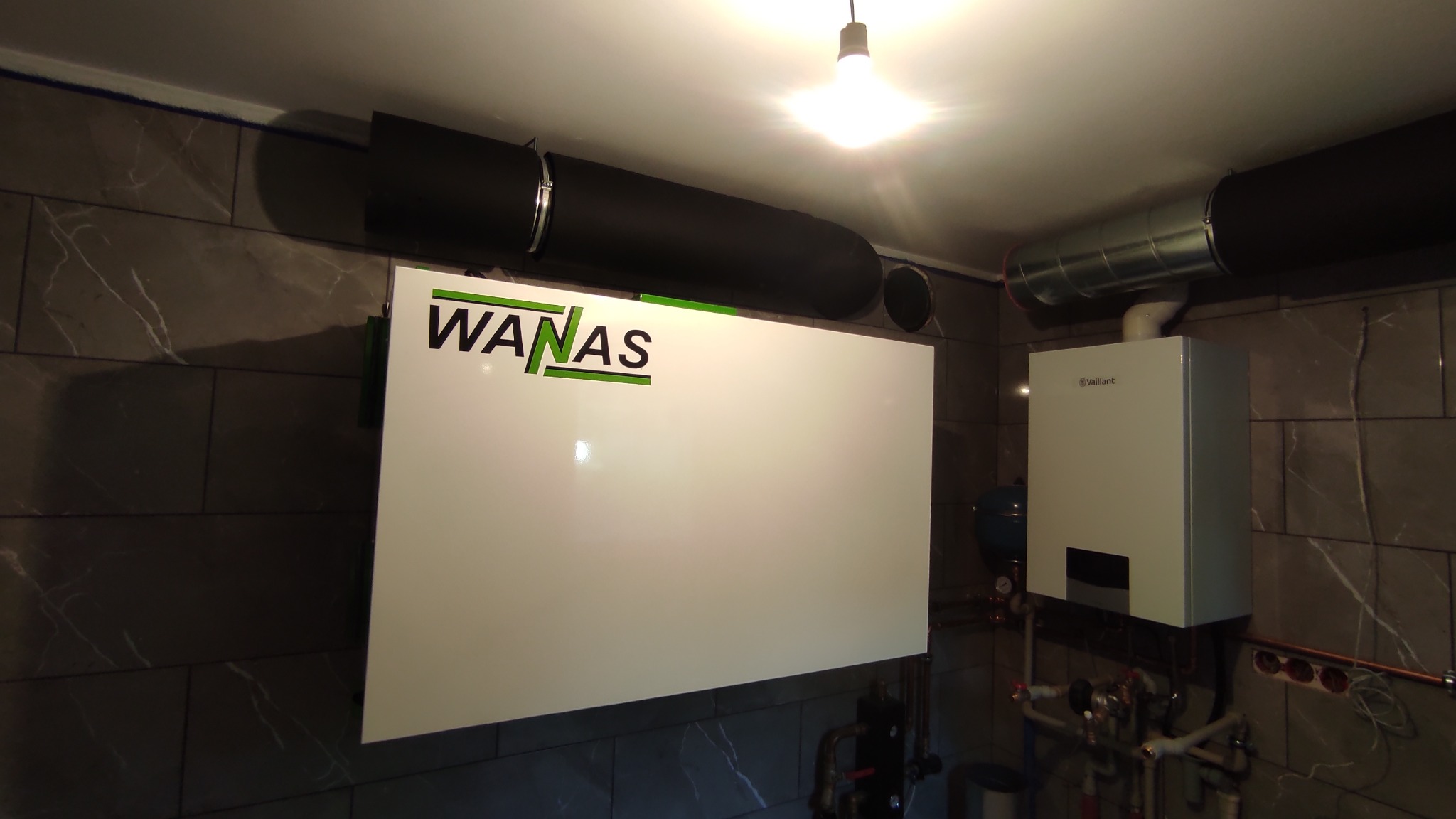Biała centrala wentylacyjna z rekuperacją marki WANAS zamontowana na ścianie z szarych płytek imitujących marmur, widoczne czarne rury wentylacyjne i piec gazowy Vaillant.