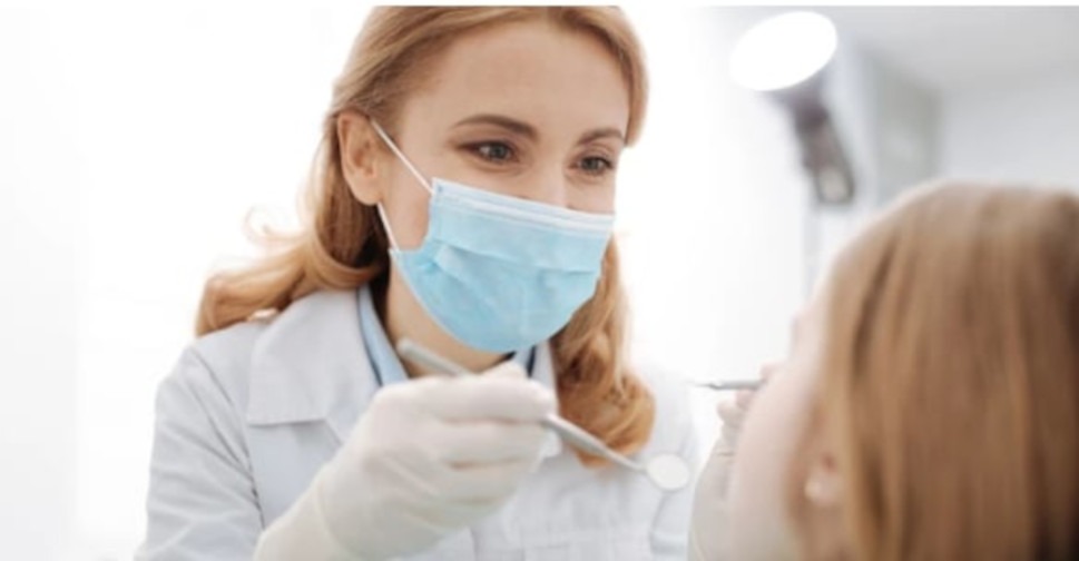 Lekarz w masce i białym kitlu, delikatnie pochylony nad pacjentem, używa narzędzi dentystycznych podczas zabiegu, jasne oświetlenie