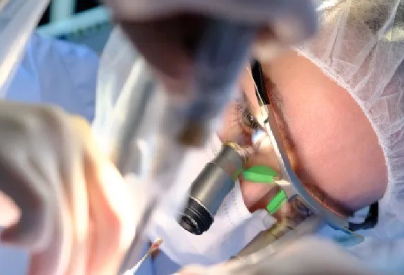 Pacjent podczas zabiegu stomatologicznego z użyciem mikroskopu, widoczne zielone paski ochronne i narzędzia w dłoniach lekarza.