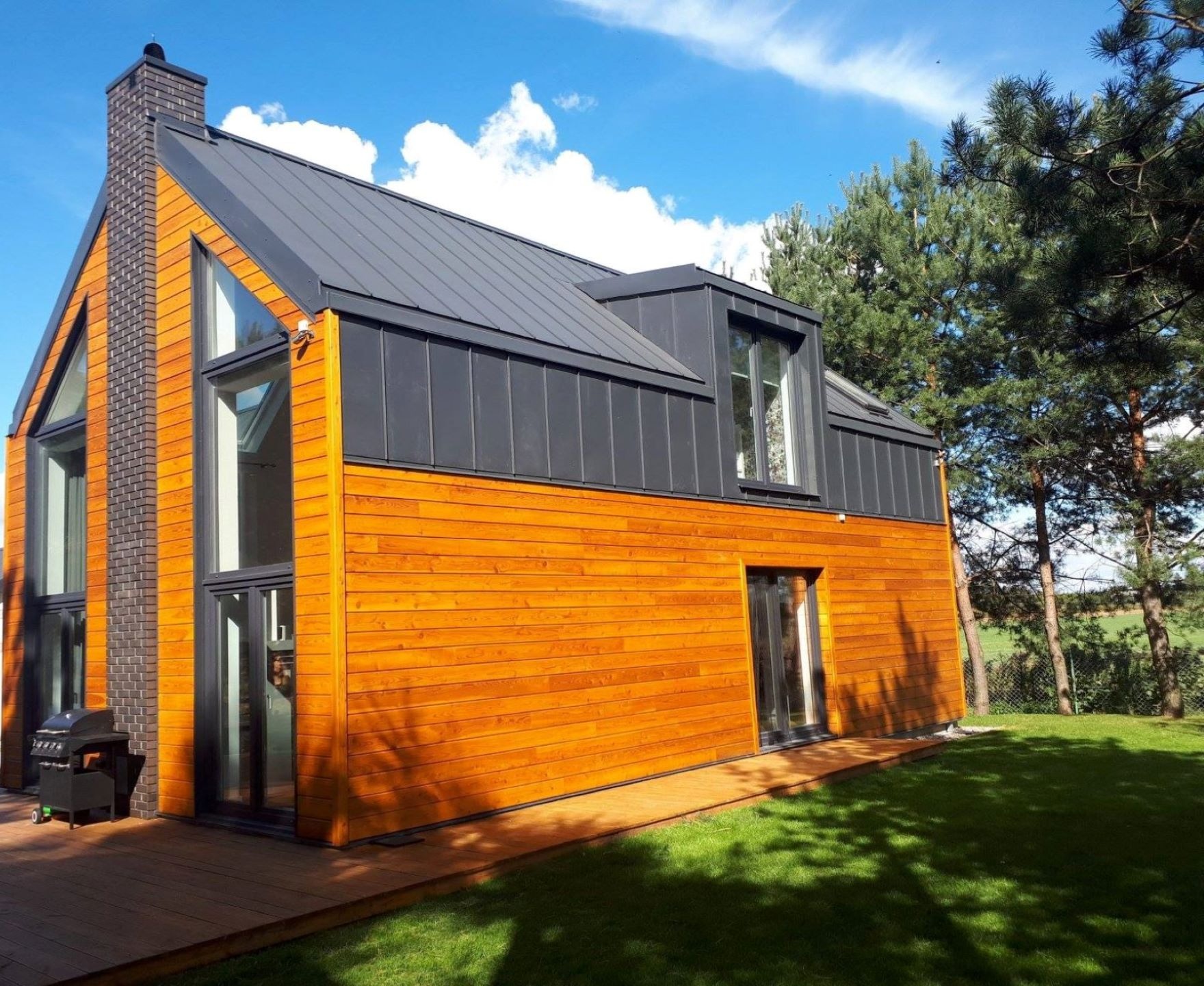 Nowoczesny dom z drewnianą elewacją w odcieniach brązu, czarnym dachem i kominem z cegły, otoczony zielonym trawnikiem i drzewami iglastymi, z grillem na tarasie.