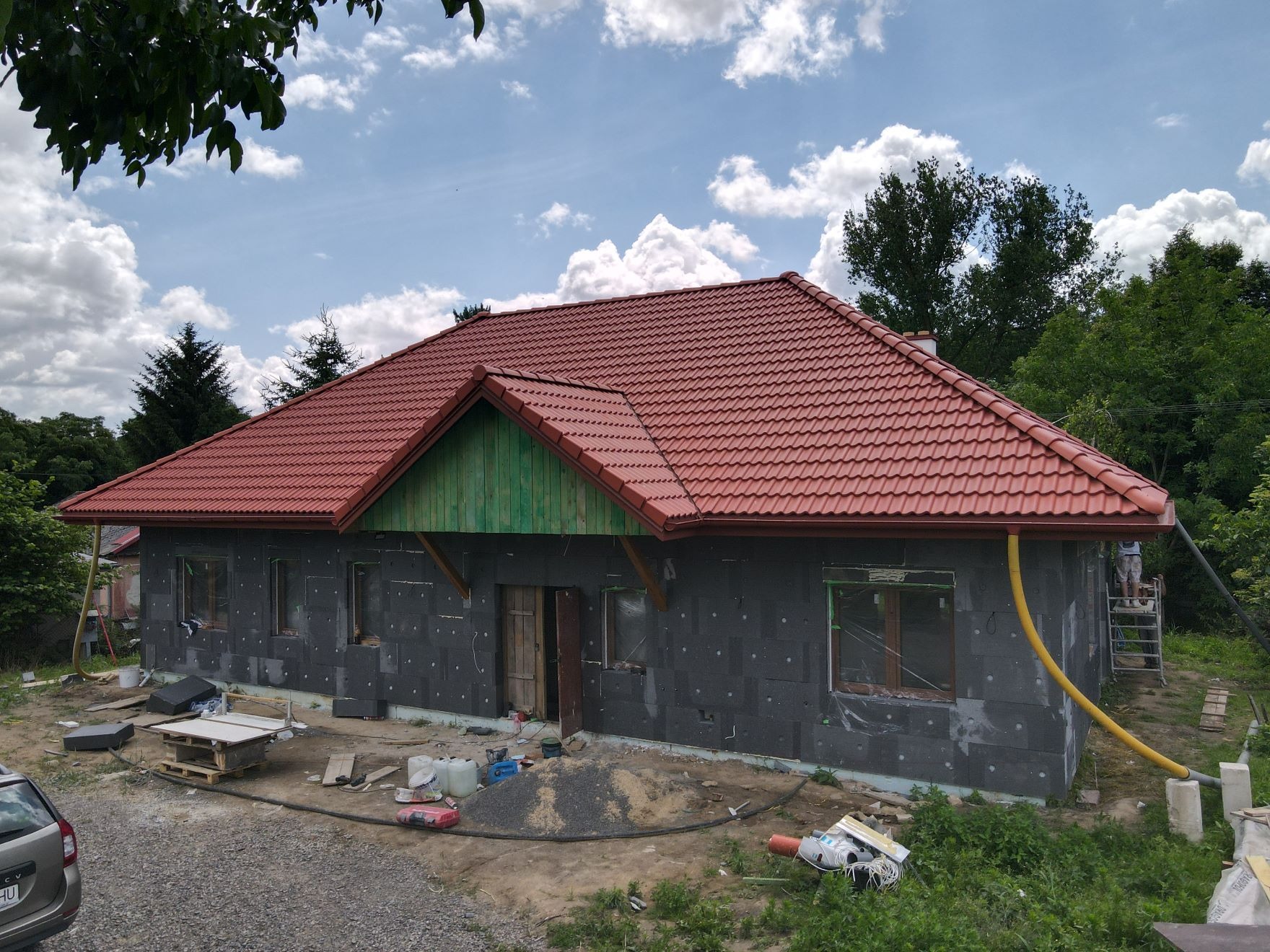 Budowa domu jednorodzinnego z czerwoną dachówką, ocieplonego styropianem grafitowym, widok na elewację w trakcie prac wykończeniowych, z widocznym rusztowaniem i materiałami budowlanymi na posesji.