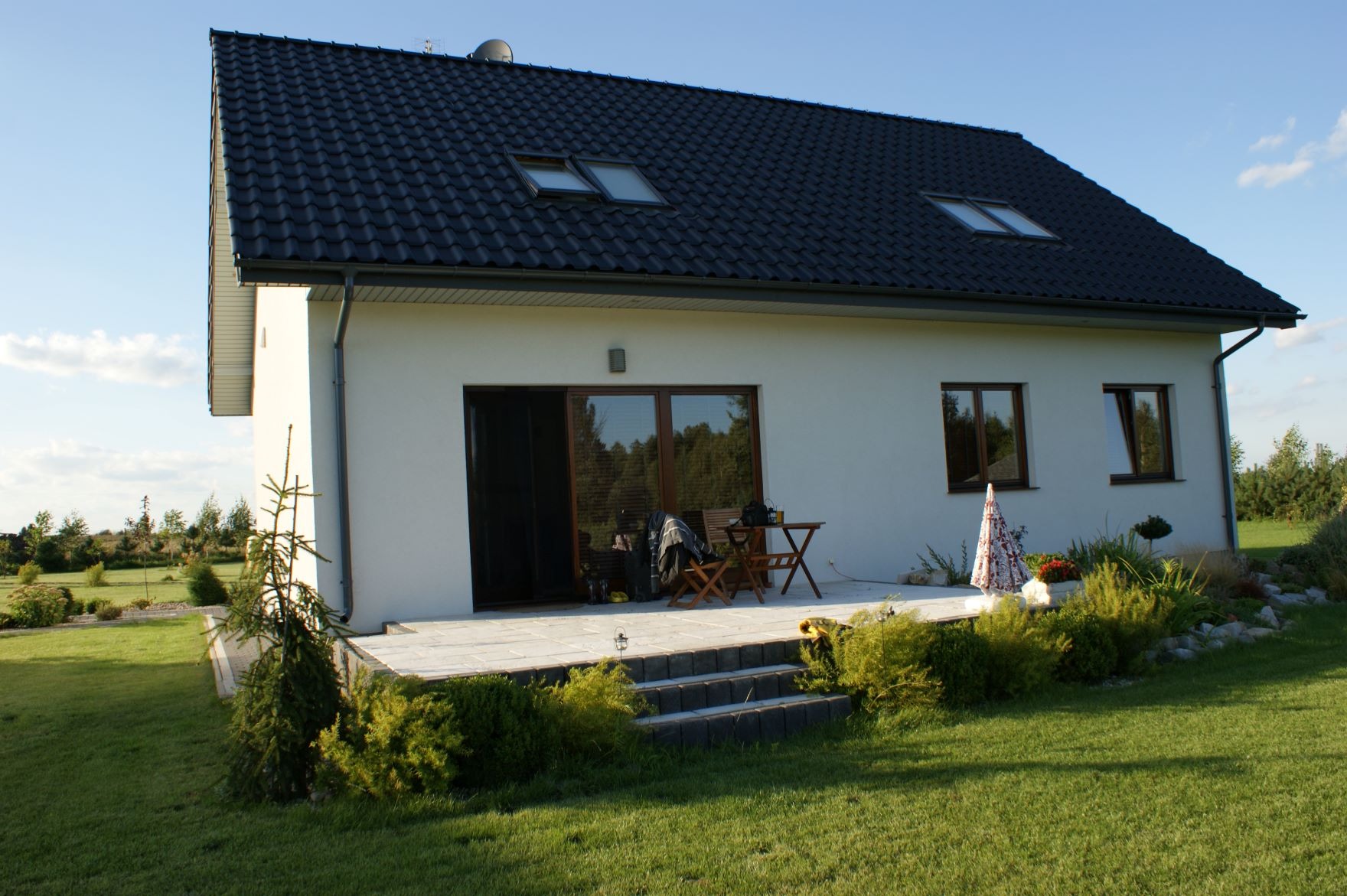 Nowoczesny dom parterowy z czarną dachówką, tarasem z meblami ogrodowymi i zadbanym trawnikiem.