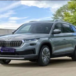 Szary SUV Škoda Kodiaq w ruchu na tle błękitnego nieba i zieleni. Widoczny przód pojazdu z charakterystycznym grillem i reflektorami.