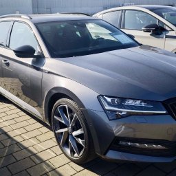 Krzysztof Witkowski - Szary samochód Skoda Superb Combi z aluminiowymi felgami, zaparkowany na brukowanej powierzchni. Widoczne inne pojazdy w tle, oświetlone jasnym światłem słonecznym.