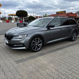 Krzysztof Witkowski - Szary samochód Skoda Superb Combi na parkingu stacji benzynowej Shell, widok z boku, pochmurne niebo w tle. Tablica rejestracyjna widoczna.