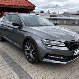 Krzysztof Witkowski - Szary samochód Skoda Superb Combi na parkingu z kostki brukowej, widok z przodu, tablica rejestracyjna NOS-64953, zadaszenie w tle.