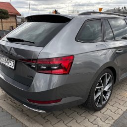 Krzysztof Witkowski - Szary samochód Skoda Superb Combi 4x4 na kostce brukowej, widok z tyłu, mokry po deszczu. Widoczne światła, tablica rejestracyjna i felgi. Pochmurne niebo w tle.