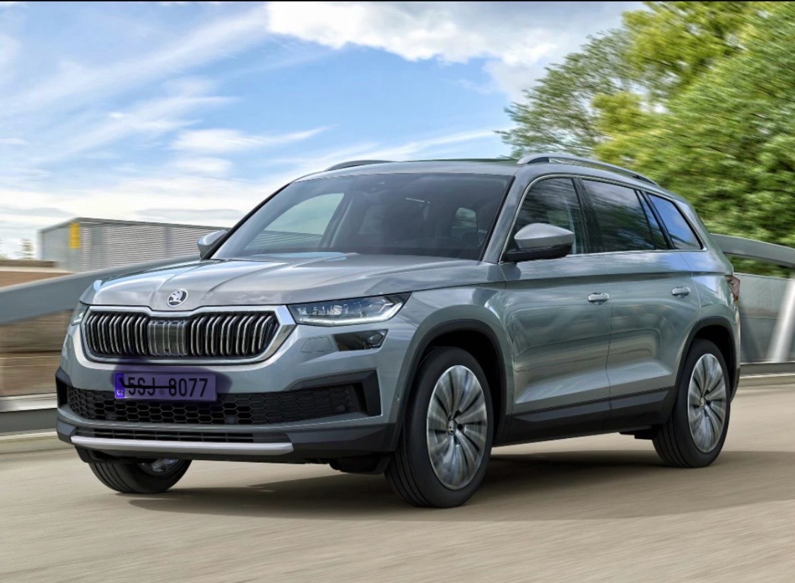 Szary SUV Škoda Kodiaq w ruchu na tle błękitnego nieba i zieleni. Widoczny przód pojazdu z charakterystycznym grillem i reflektorami.