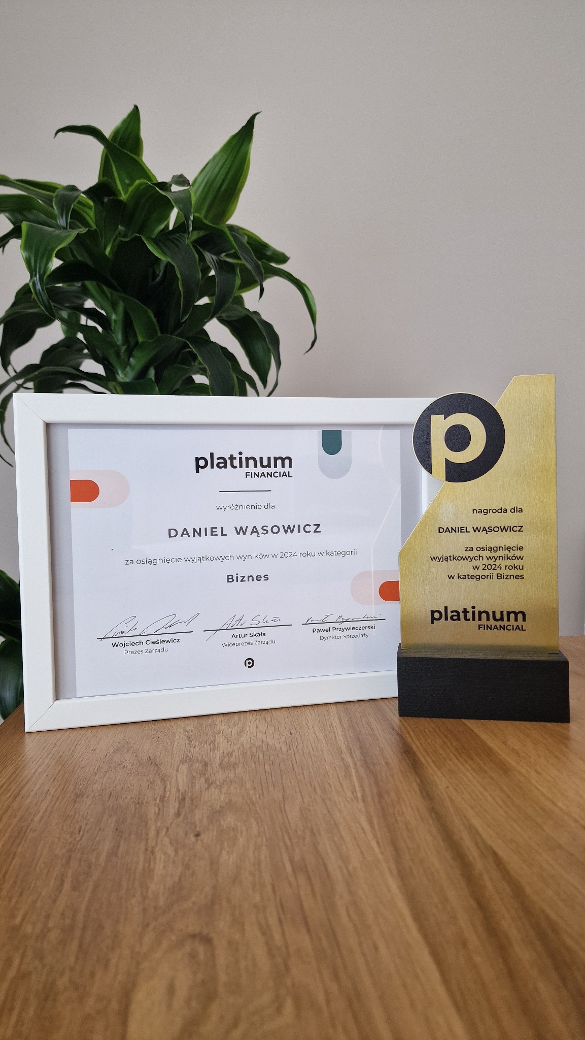 Nagroda i certyfikat Platinum Financial dla Daniela Wąsowicza za wyjątkowe wyniki w biznesie w 2024 roku, na drewnianym biurku z rośliną w tle.