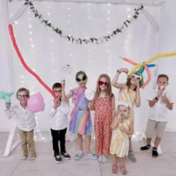 Grupa dzieci w zabawnych okularach i z balonami pozuje na tle dekoracji z lampek i kwiatów, prawdopodobnie podczas imprezy urodzinowej.