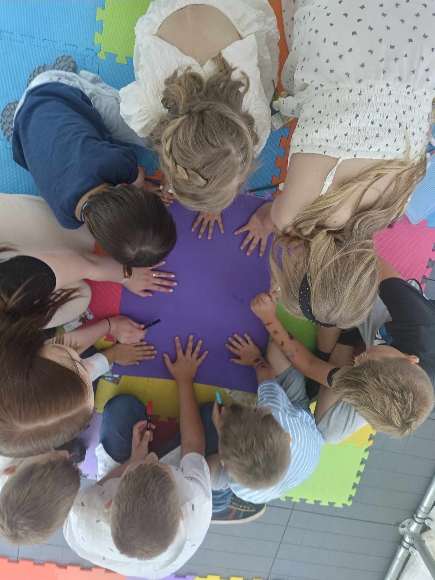 Grupa dzieci i dorosłych, widok z góry, rysuje markerami na fioletowym arkuszu papieru leżącym na kolorowej macie piankowej.