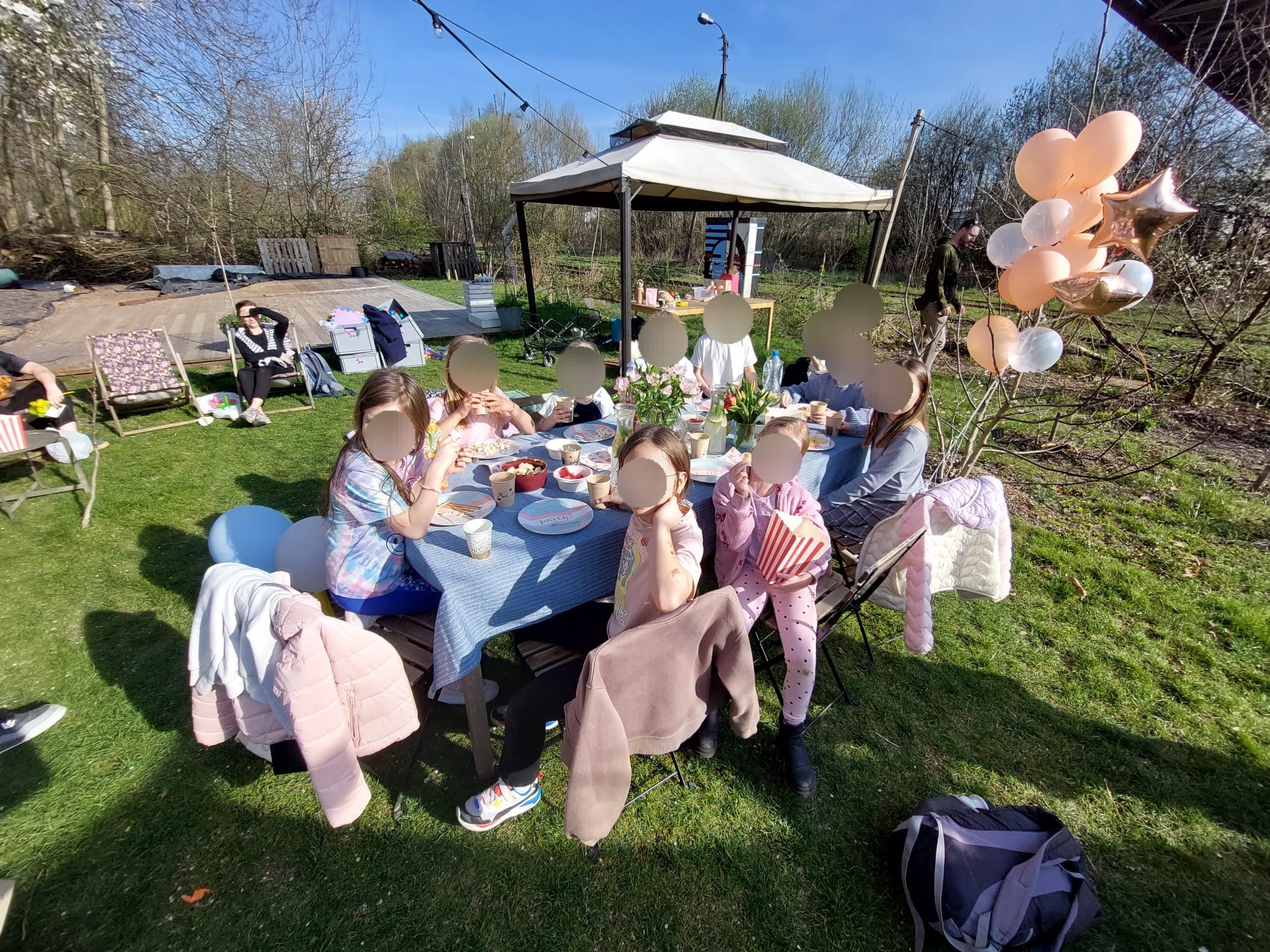 Uroczystość na świeżym powietrzu: dzieci siedzą przy stole z niebieskim obrusem, dekoracje z balonów, w tle altana i zieleń. Słoneczny dzień.