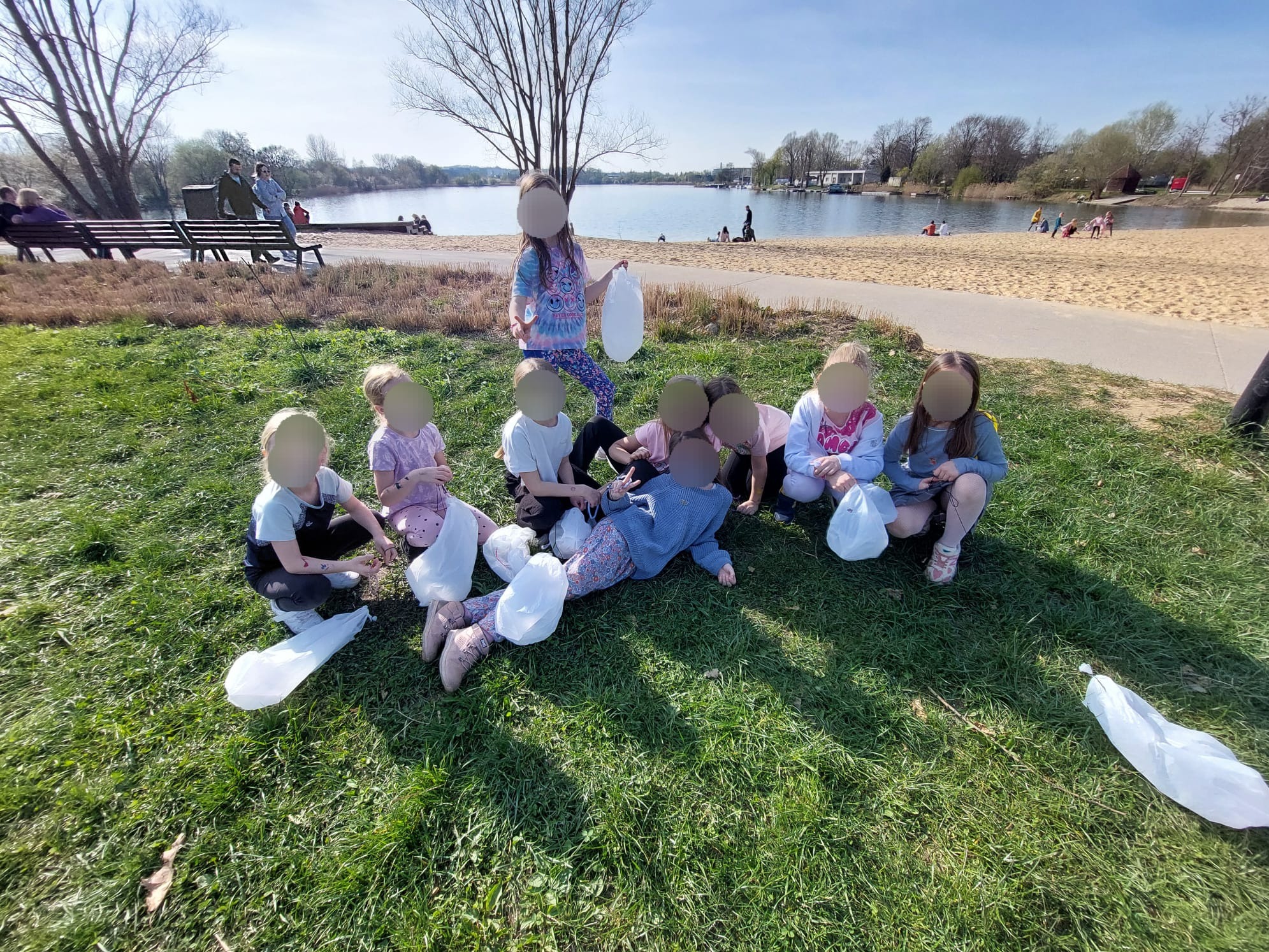 Grupa dzieci zbiera śmieci w workach na trawie nad jeziorem, w tle plaża i spacerujący ludzie. Akcja sprzątania zorganizowana przez animatora dla dzieci.