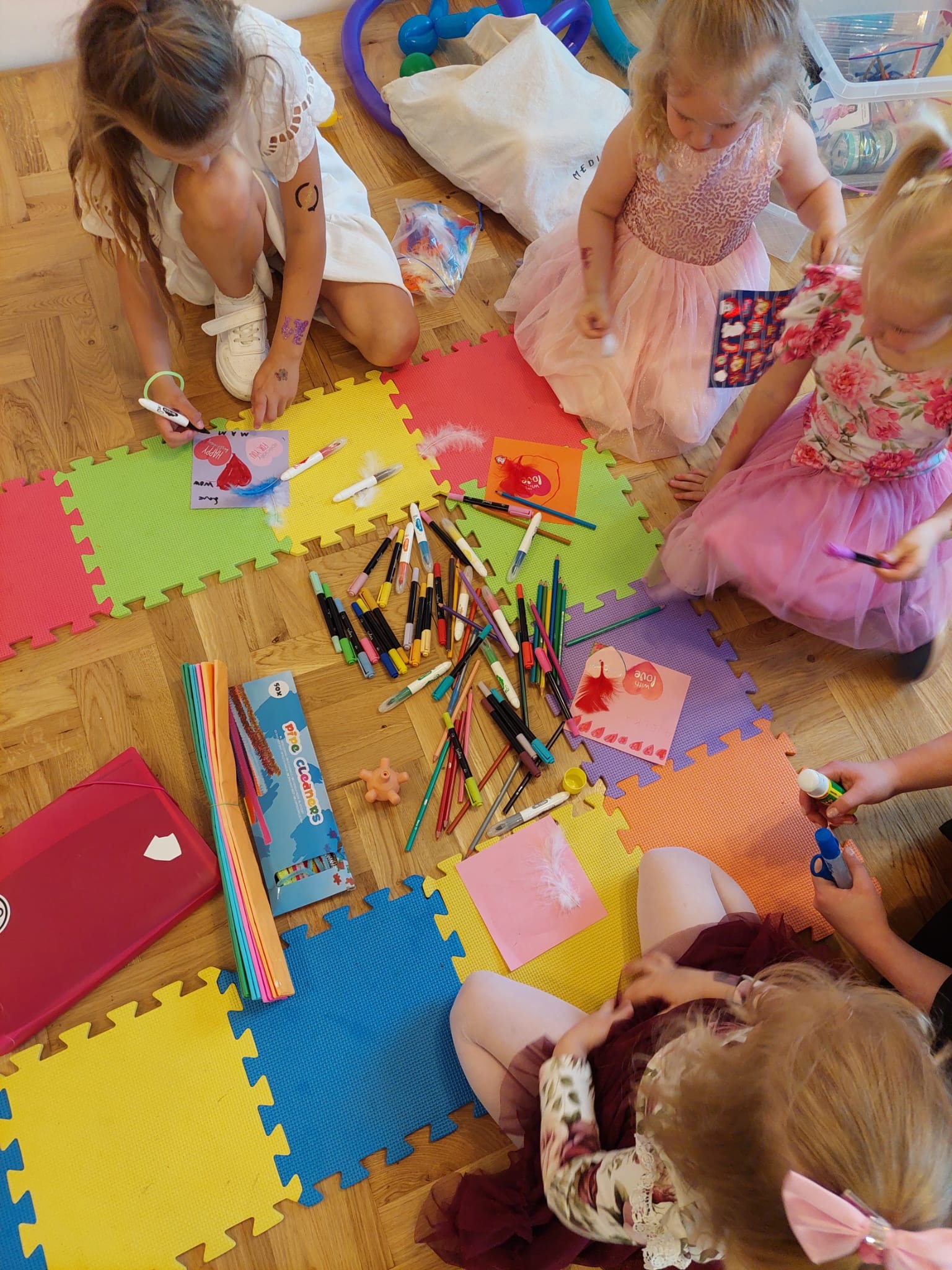 Dzieci w tiulowych sukienkach tworzą kartki okolicznościowe, siedząc na kolorowych puzzlach podłogowych. Widoczne pisaki, piórka i inne materiały plastyczne.