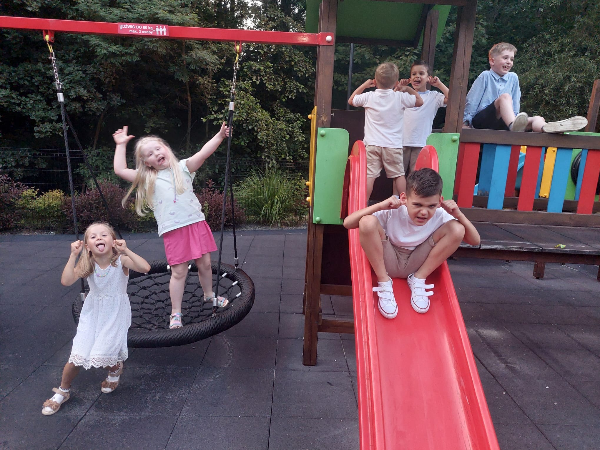 Rozbawione dzieci na placu zabaw: huśtawka typu 'gniazdo', zjeżdżalnia i drewniana konstrukcja. Radosne miny i gesty, zabawa na świeżym powietrzu.