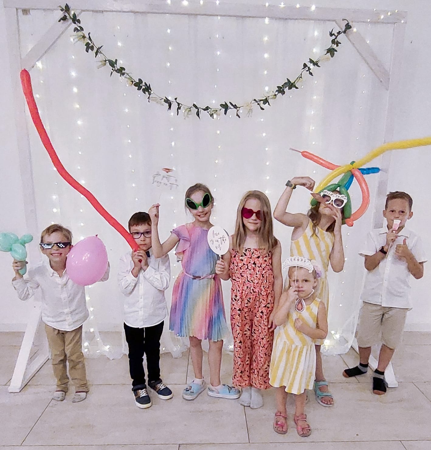 Grupa dzieci w zabawnych okularach i z balonami pozuje na tle dekoracji z lampek i kwiatów, prawdopodobnie podczas imprezy urodzinowej.