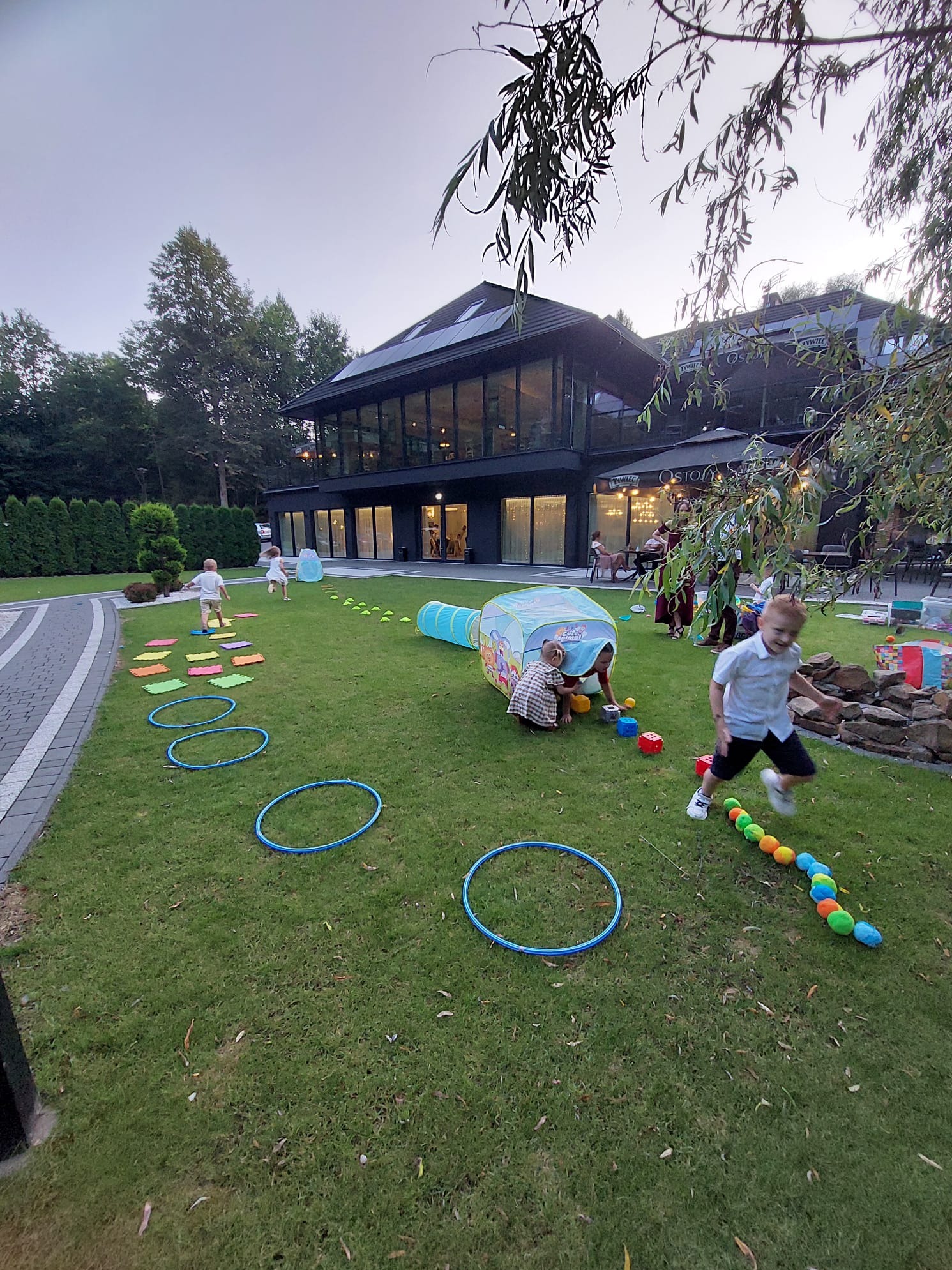 Animator prowadzi gry na trawie: bieg z piłkami, tunel, hula-hop. Dzieci bawią się na zewnątrz, w tle nowoczesny budynek z dużymi oknami i zielony trawnik.