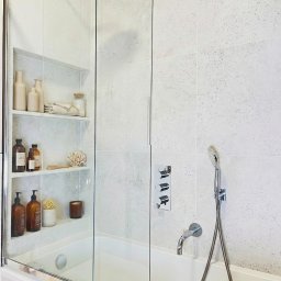 Harmony Ewa Jaworska - Jasna łazienka z wanną za szklanym parawanem, półki z kosmetykami w ścianie, armatura w chromie, płytki Moonrow white. Minimalistyczny design.