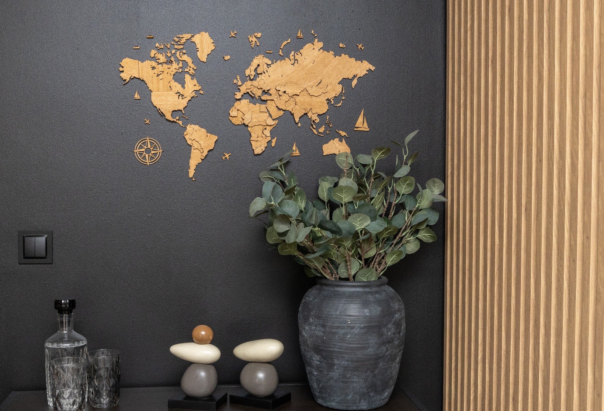 Dekoracja ściany z drewnianą mapą świata i panelem ściennym w Suchym Dworze. Wazon z eukaliptusem i szklane naczynia na półce dodają wnętrzu charakteru.