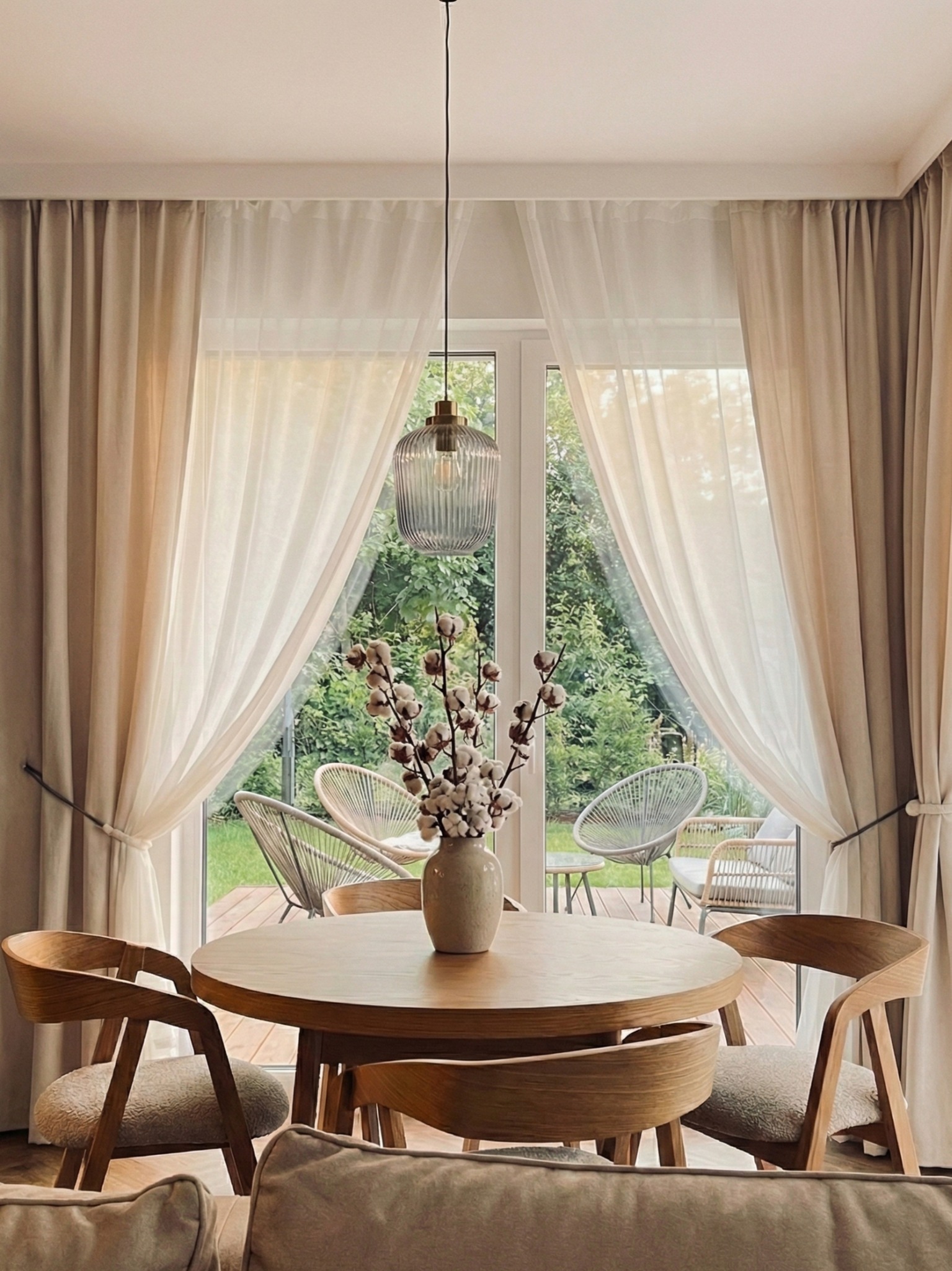 Jasne wnętrze jadalni z okrągłym stołem i krzesłami z drewna. W tle okno z widokiem na taras i ogród, zasłonięte delikatnymi, beżowymi zasłonami.