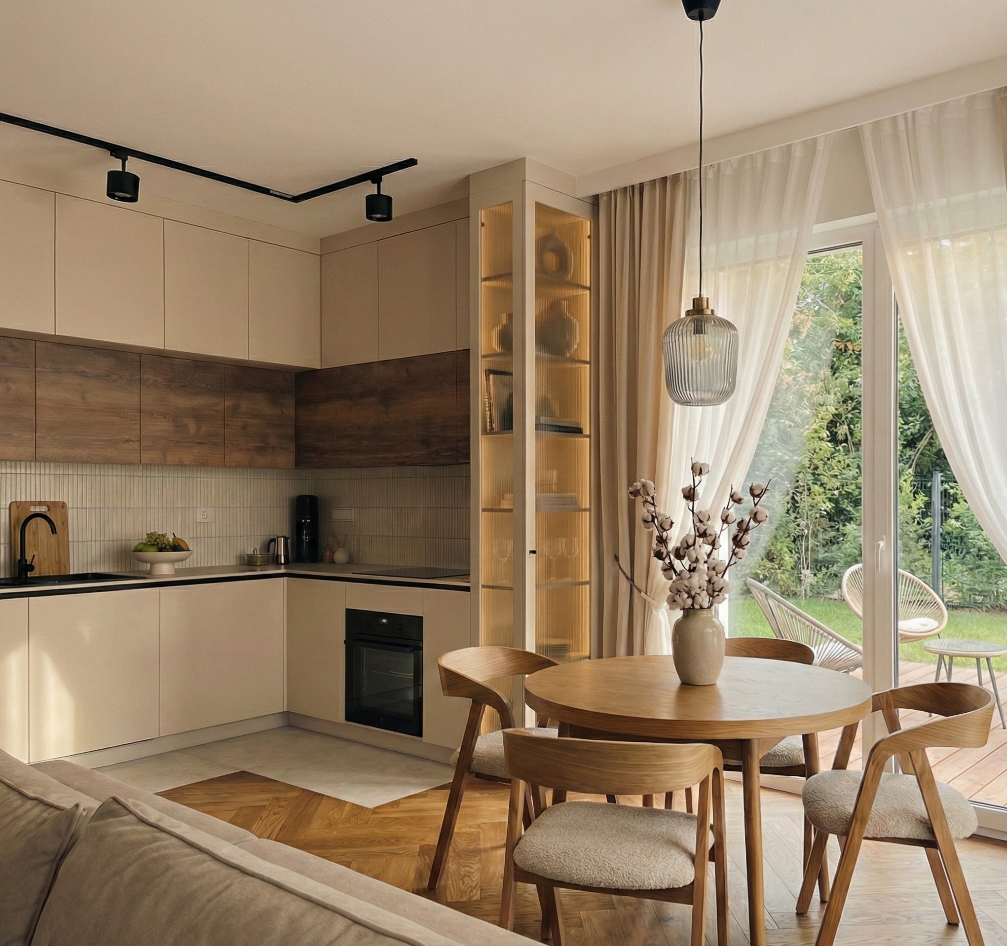 Jasna kuchnia z drewnianymi akcentami, okrągły stół jadalny z krzesłami, przeszklona witryna z podświetleniem, widok na ogród przez okno balkonowe.
