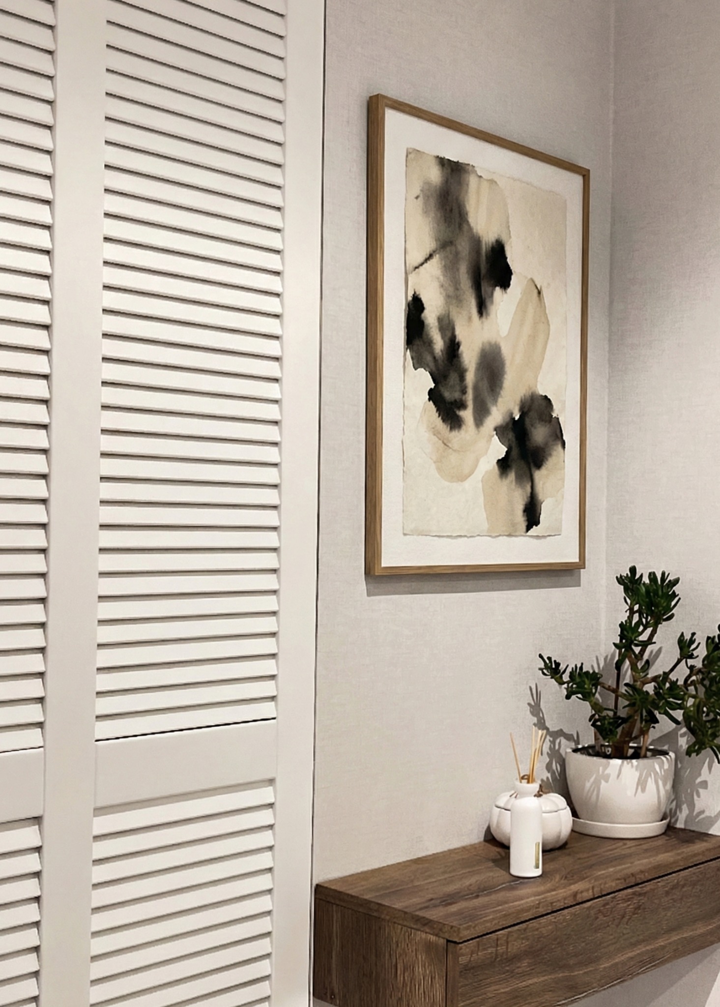 Minimalistyczny korytarz: białe żaluzjowe drzwi, abstrakcyjny obraz w drewnianej ramie, drewniana konsola z rośliną w doniczce i dyfuzorem zapachowym.