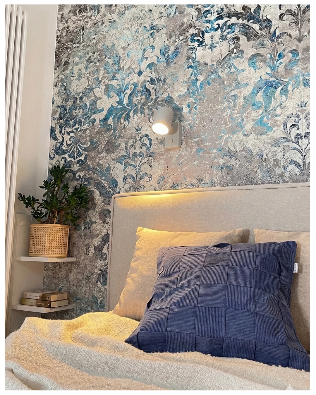 Sypialnia z wzorzystą tapetą w odcieniach szarości i błękitu, oświetlona kinkietem. Na łóżku dekoracyjna, pleciona poduszka w kolorze granatowym.